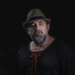 Moody portrait of a middle-aged man wearing a fedora hat, featuring a pensive expression against a dark backdrop.